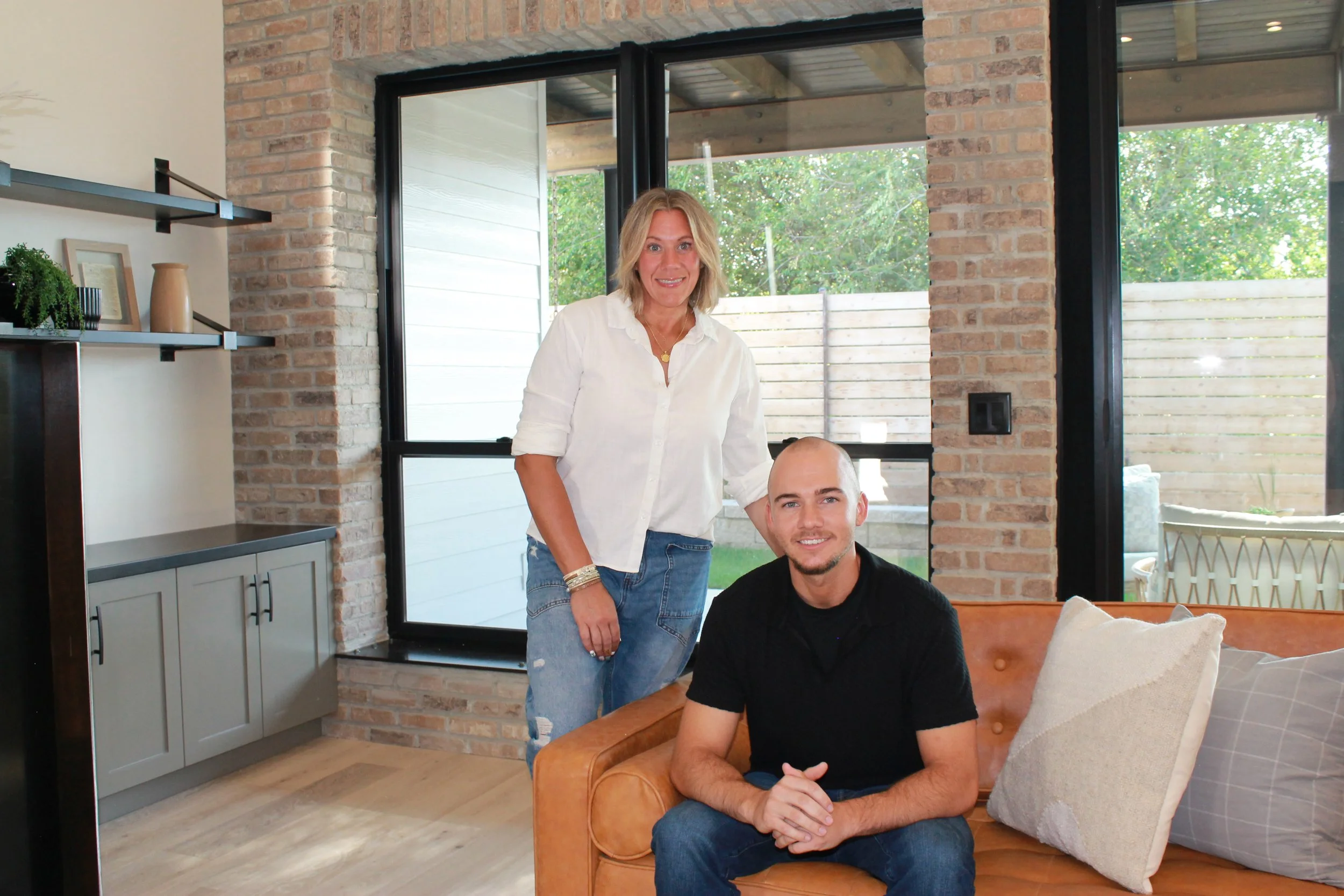 Two people, a woman and a man, inside a modern living room with large windows, brick walls, and wooden flooring. The woman is standing, smiling, wearing a white shirt and jeans. The man is sitting on a sofa, smiling, wearing a black shirt.