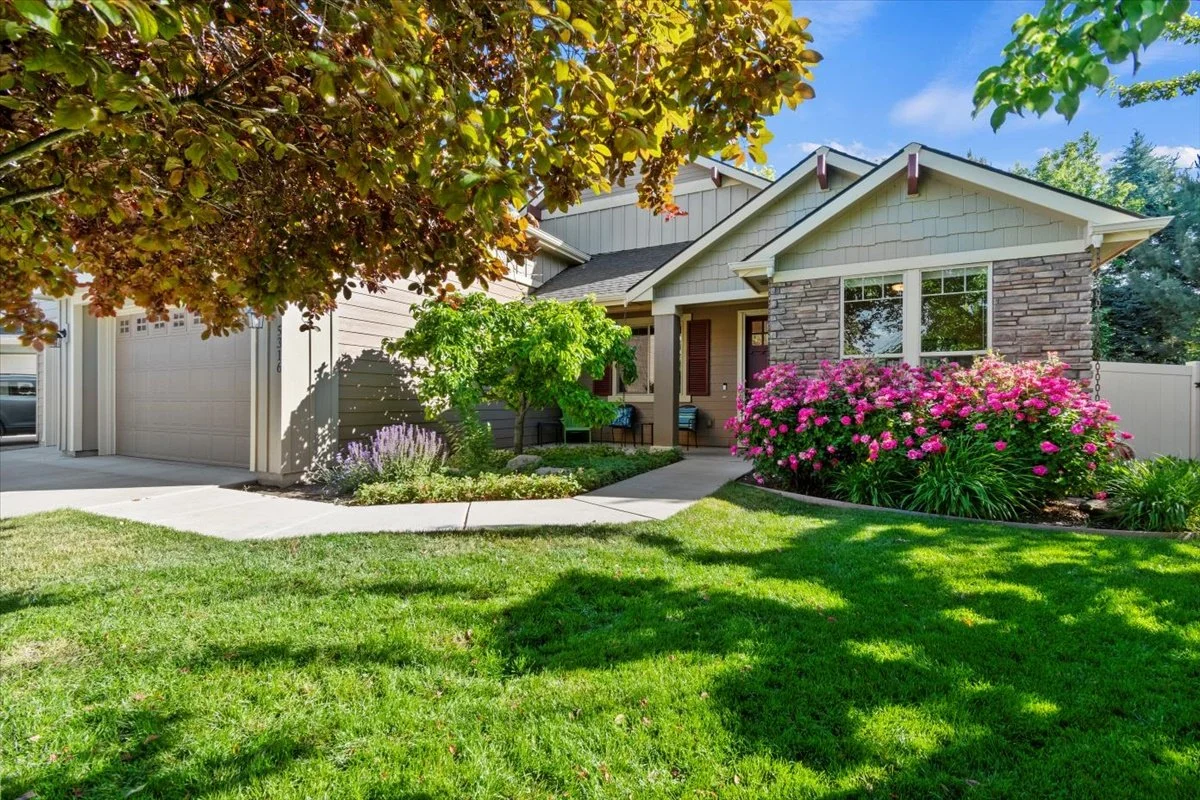 A suburban house with a well-maintained front yard, blooming pink flowers, green bushes, and a sidewalk leading to the front door.