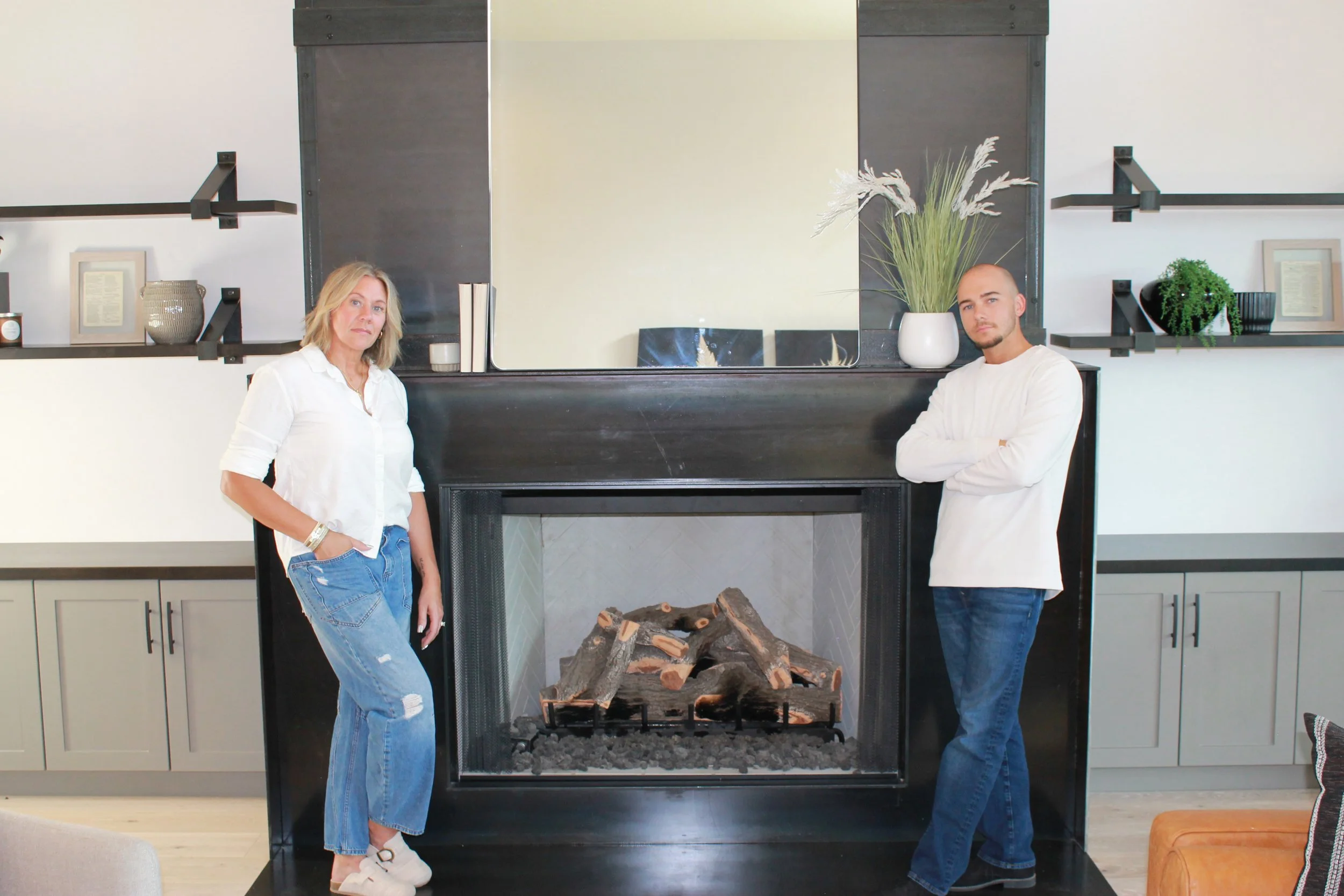A woman with shoulder-length blonde hair in a white shirt and ripped jeans stands on the left, and a man with a shaved head in a white sweater and jeans stands on the right, both posing with arms crossed in front of a modern black fireplace with a large mirror and decorative plants on the mantle in a bright living room.