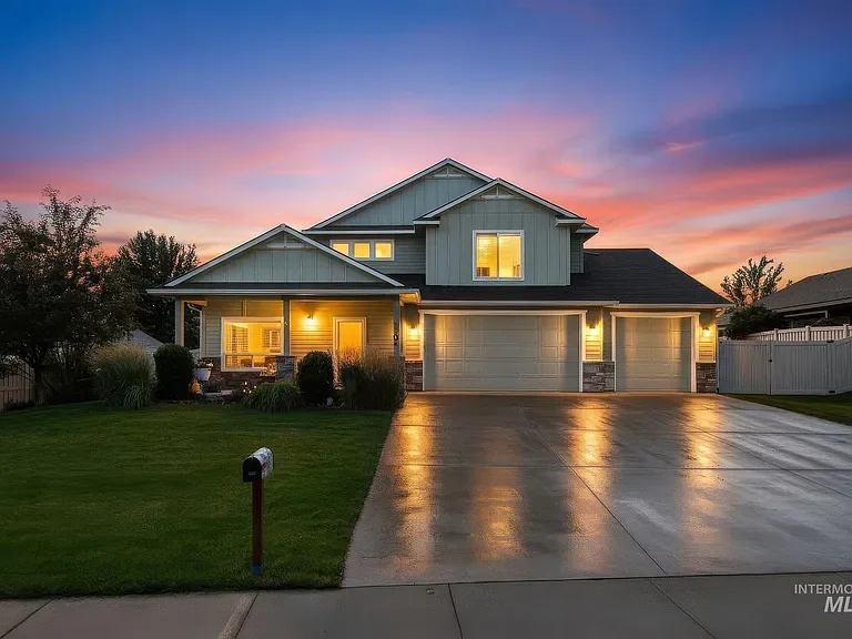 Modern two-story house at sunset with front lawn, illuminated windows, and wet driveway.