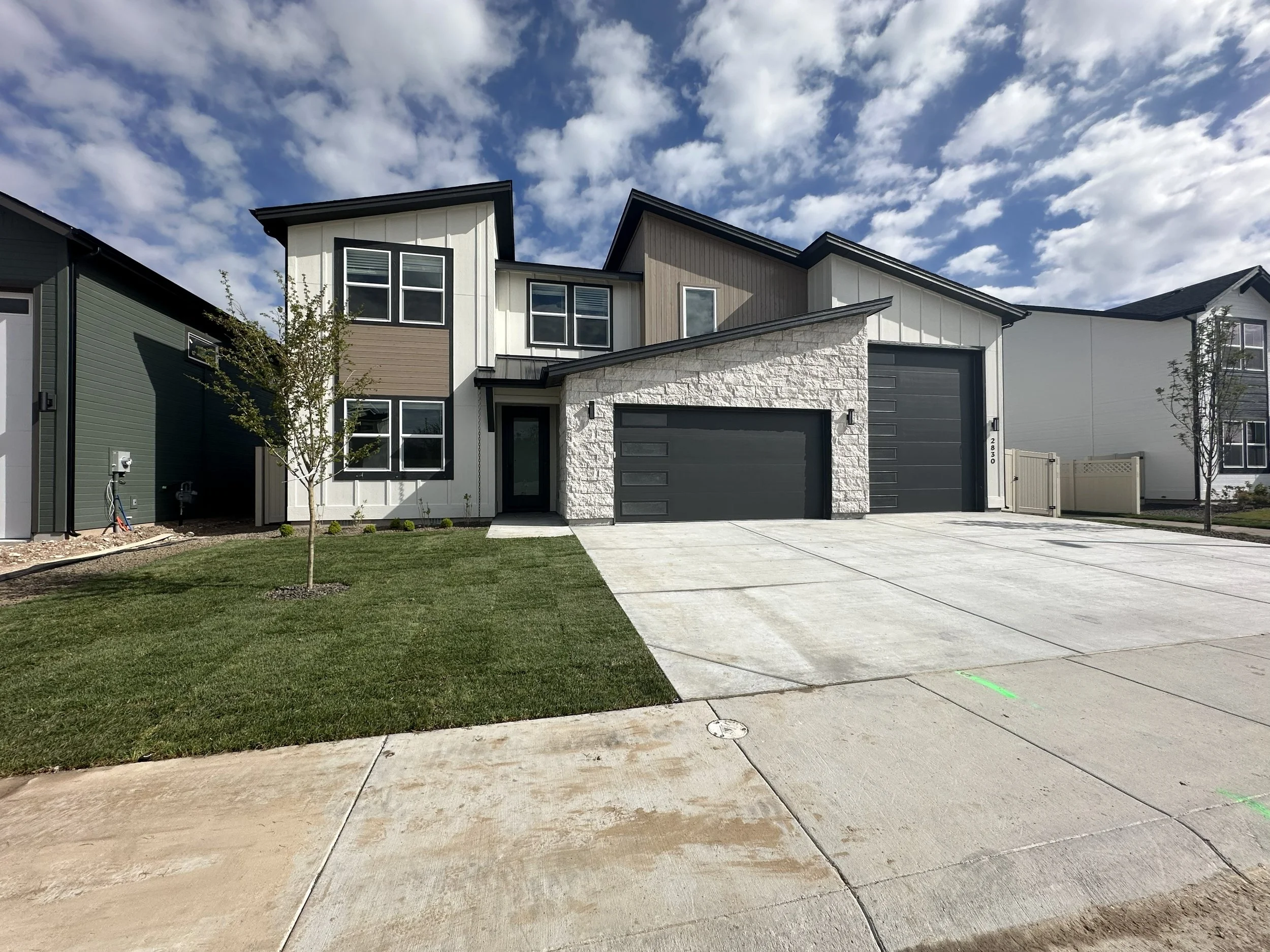 View of modern multiple-story residential houses with garage doors, a concrete driveway, small trees, and a sky with scattered clouds.