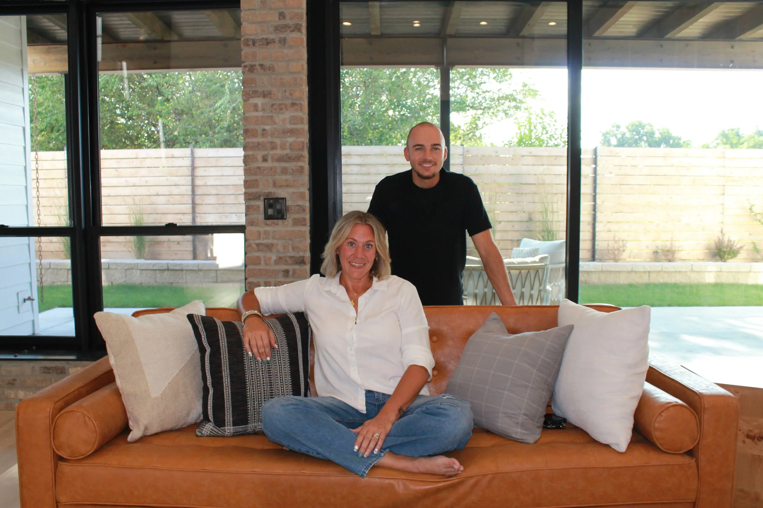 A woman with blonde hair sitting on a tan leather sofa, smiling, with her legs crossed and resting her hand on her knee. A man with a shaved head and beard standing behind her, also smiling. The sofa has several pillows, and the background features large glass windows with a view of a backyard with a wooden fence and trees.