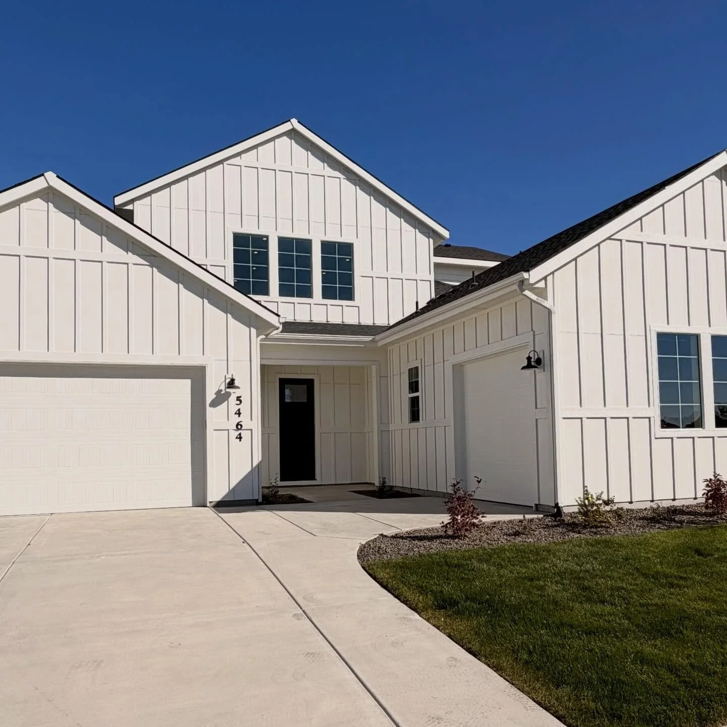 Front view of a modern white house with multiple gable roofs, a two-car garage, and a small landscaped yard under a clear blue sky.