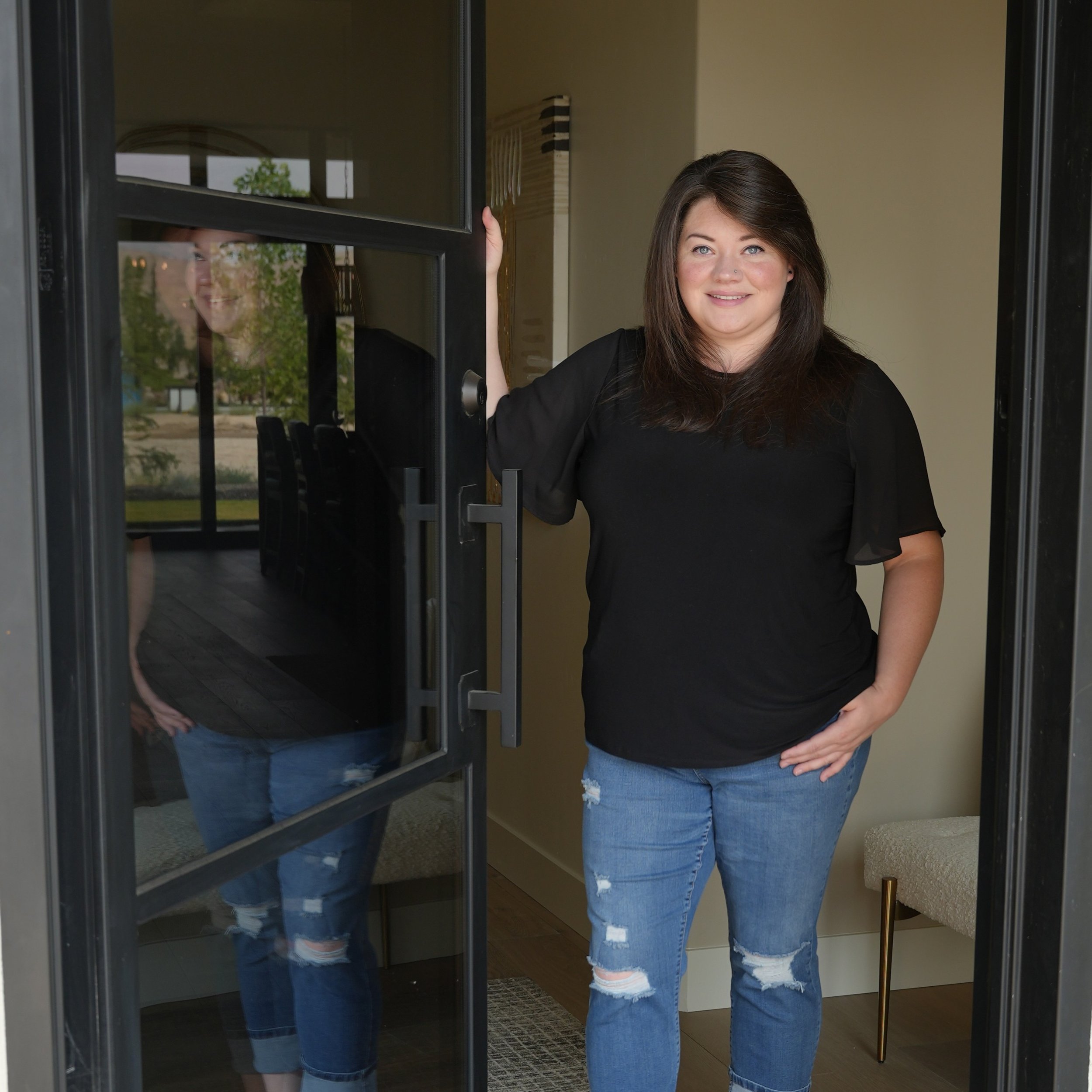 A woman with brown hair wearing a black shirt and ripped jeans stands at an open front door, smiling and holding the door.