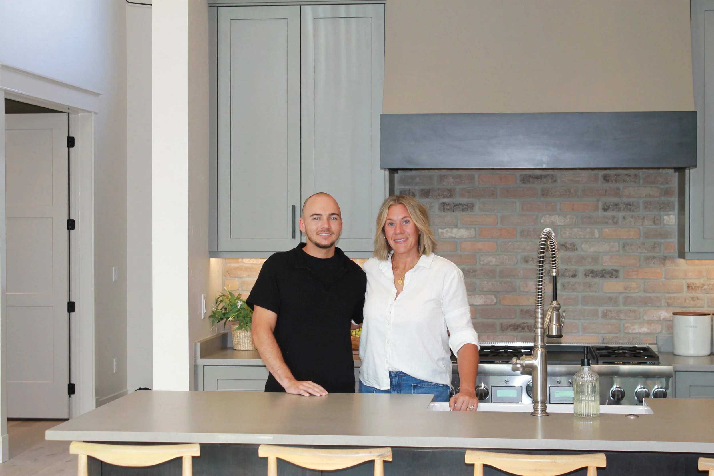 A man and woman standing together in a kitchen, smiling at the camera. The kitchen has a brick backsplash, a gray cabinet, and a sink with a modern faucet.