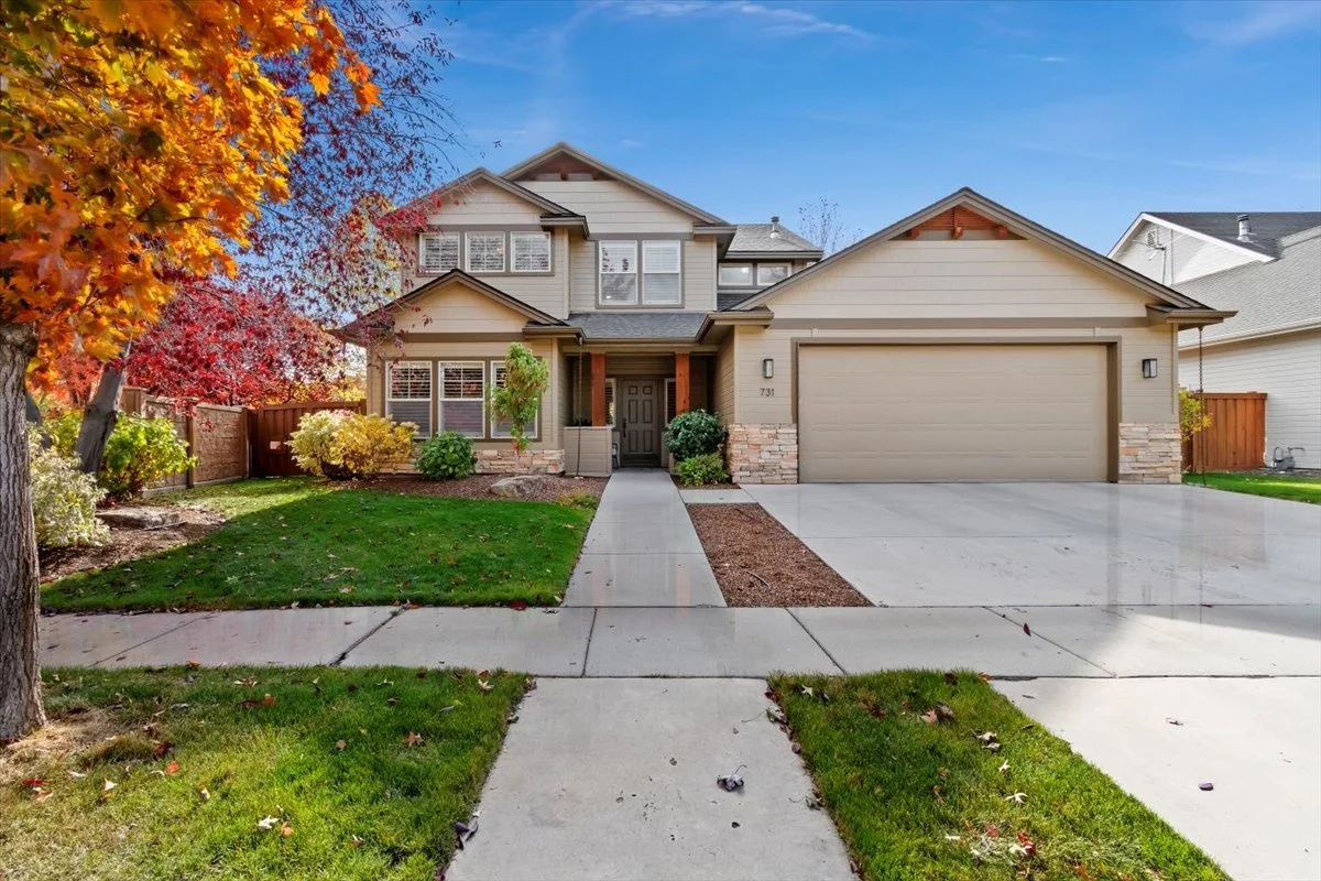 A modern two-story house with beige siding, a two-car garage, a concrete driveway, and a front porch, surrounded by trees with autumn-colored leaves and a well-manicured lawn.