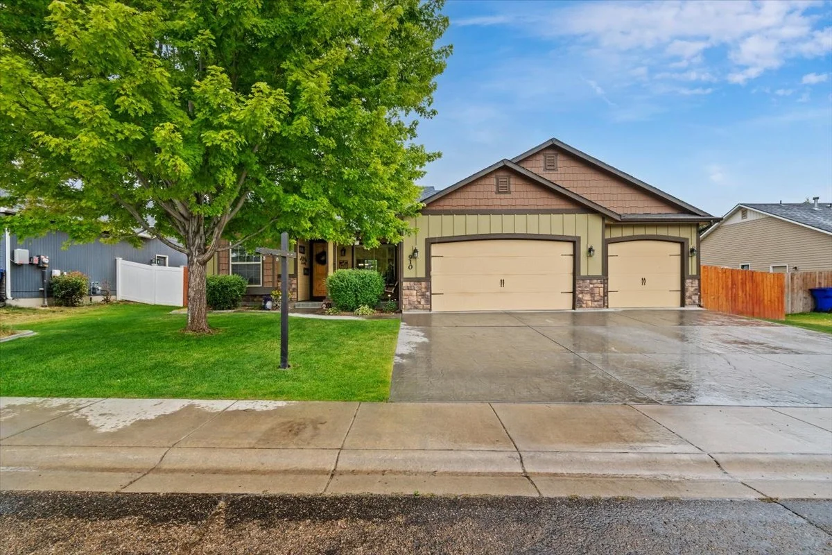 A house with a large green tree in the front yard, two-car garage, and a wet concrete driveway, after rain under a blue sky with some clouds.