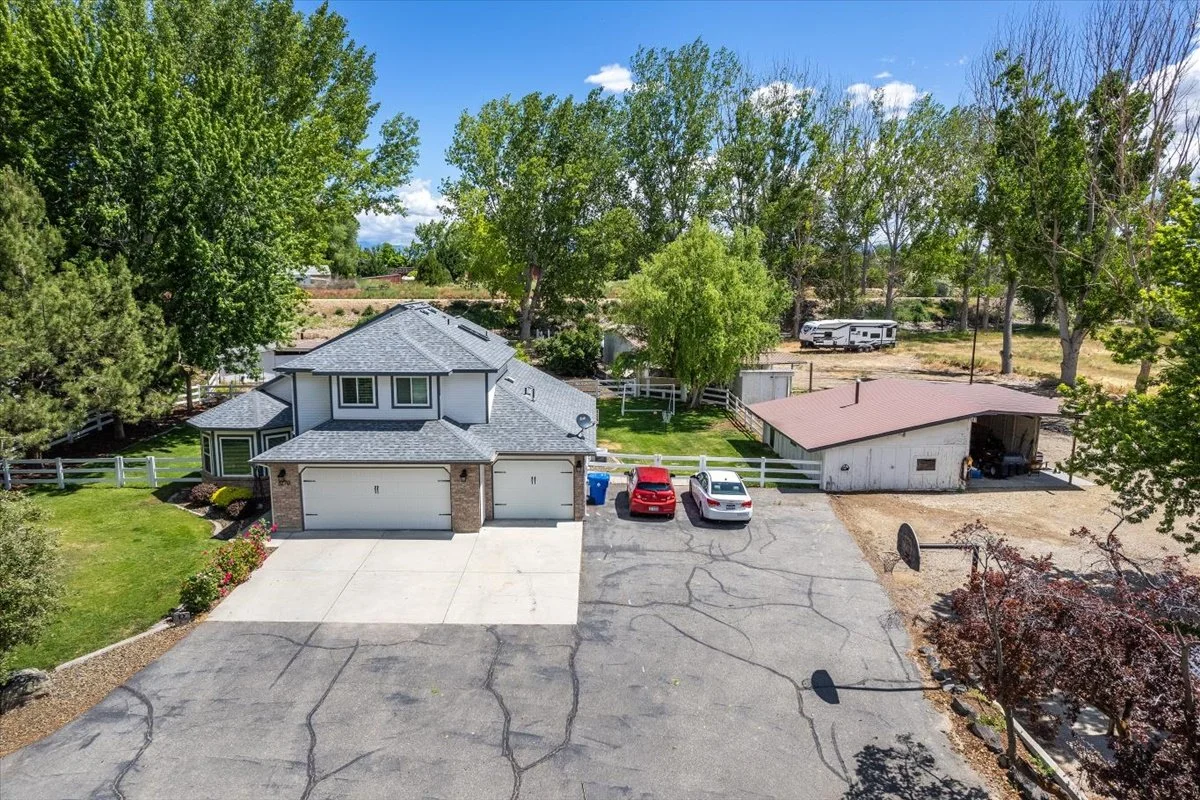 Aerial view of a two-story house with attached garage, a small shed, and parked cars in the driveway, surrounded by green trees and open land.