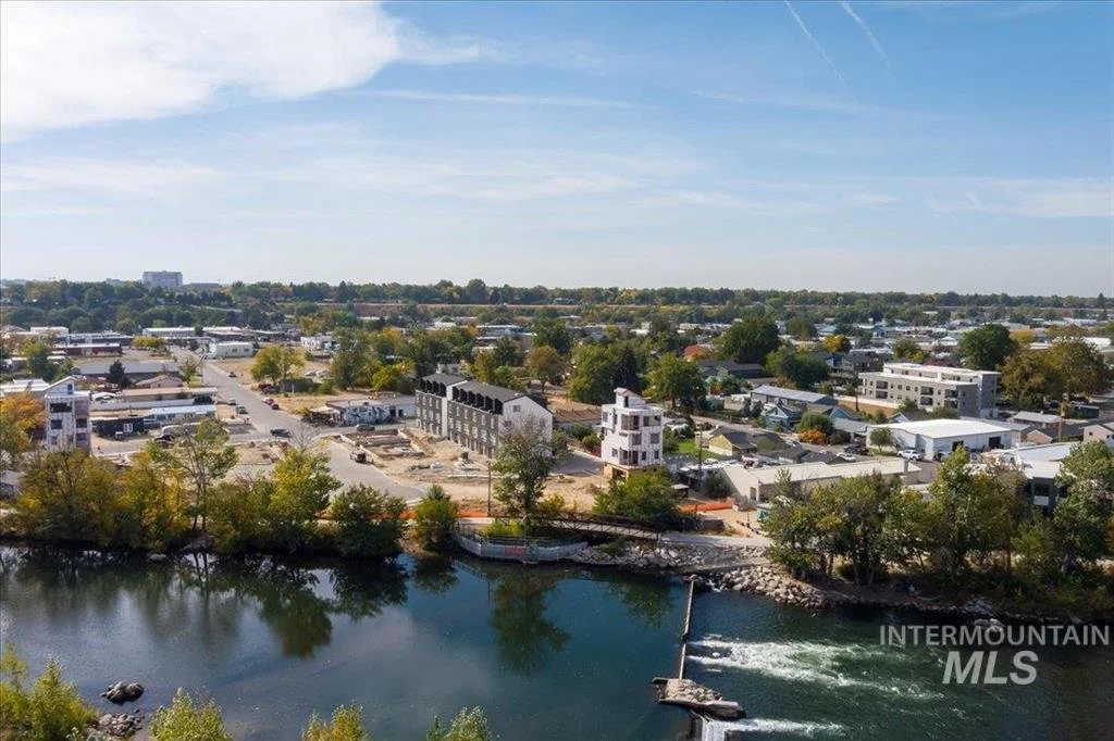 Aerial view of a cityscape with a river in the foreground, buildings, trees, and roads under a partly cloudy sky.