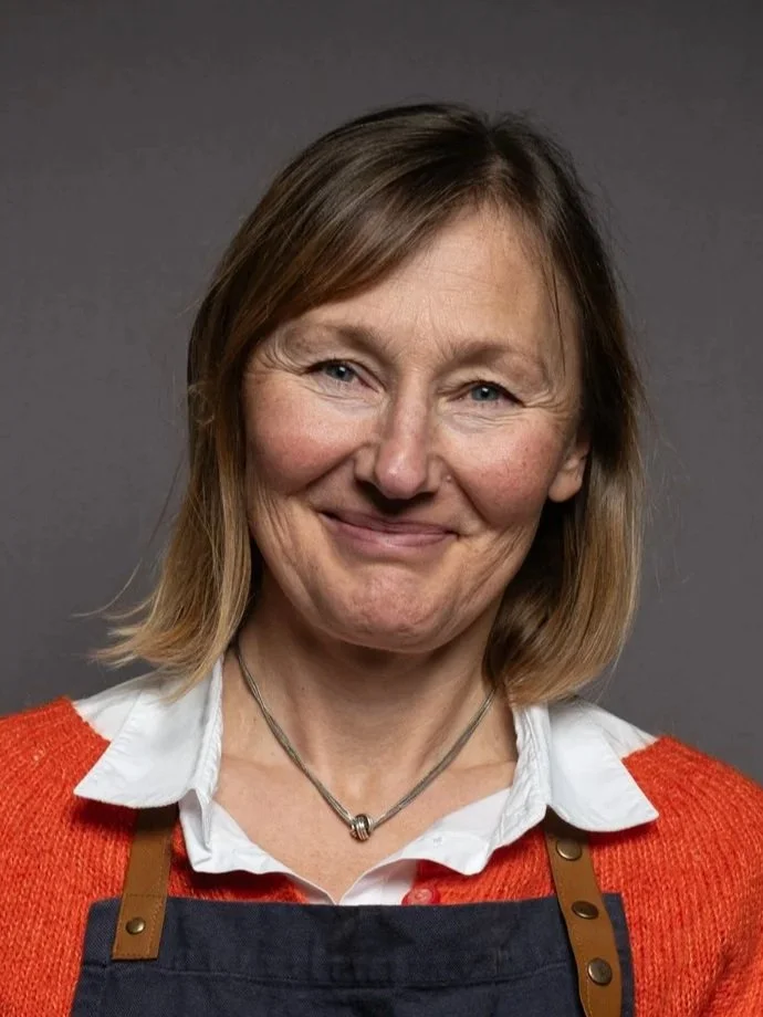 Kvinne med skulderlangt brunt hår, iført en hvit skjorte, rød genser og en mørk forklæde, smilende mot kamera. Grå bakgrunn.