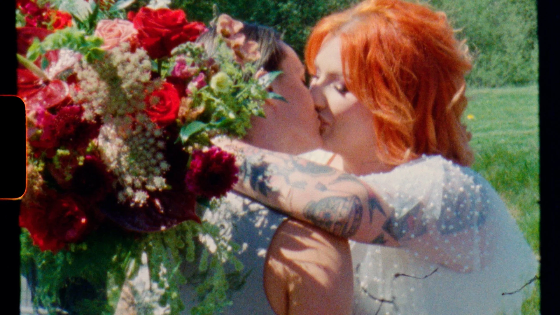Lesbian wedding, two women one with red hair and tattoos, kissing outdoors near a large bouquet of colorful flowers. Captured on Super 8 film.