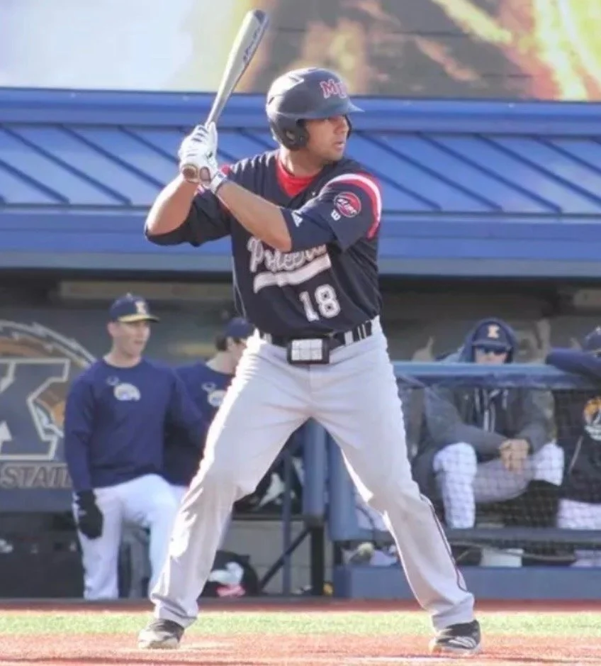 A baseball player in a black and red uniform with the number 18 on it, standing at home plate and preparing to swing a bat. The player is wearing a helmet and gloves. In the background, there are teammates and spectators watching the game.