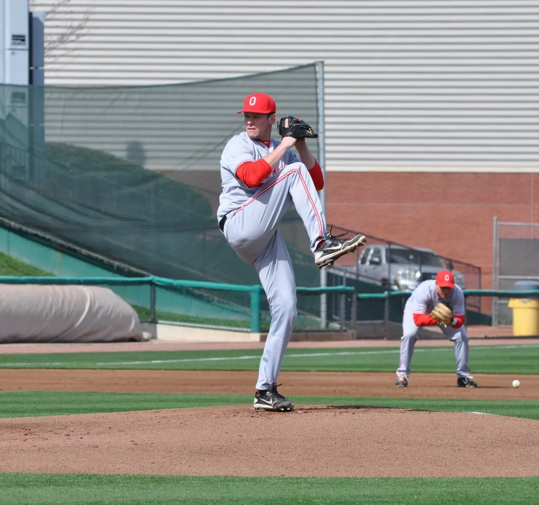 Young male baseball pitcher in a gray and red uniform preparing to throw a pitch on a baseball field with teammates in the background.