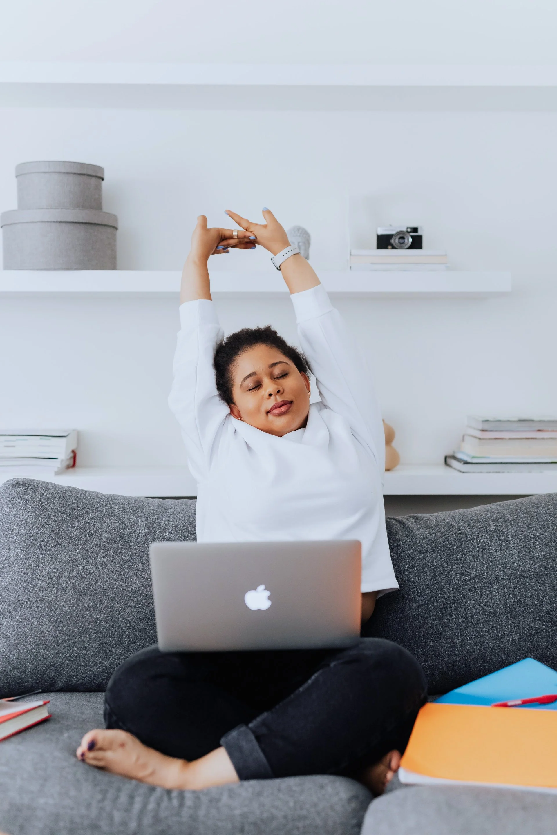 woman in white sweatshirt, sitting on couch, stretching her arms up with laptop on lap