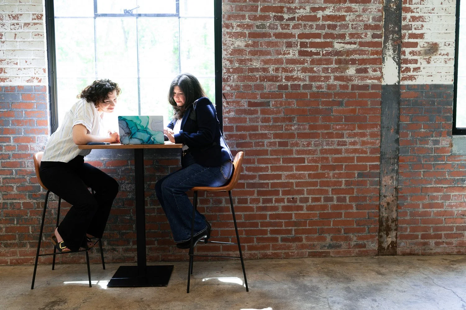Carolyn Leasure, website designer and developer at Maypop Creative Studio, sits with a client reviewing Squarespace website conversion for her service provider site, in tall chairs in front of a large window and weathered brick wall.