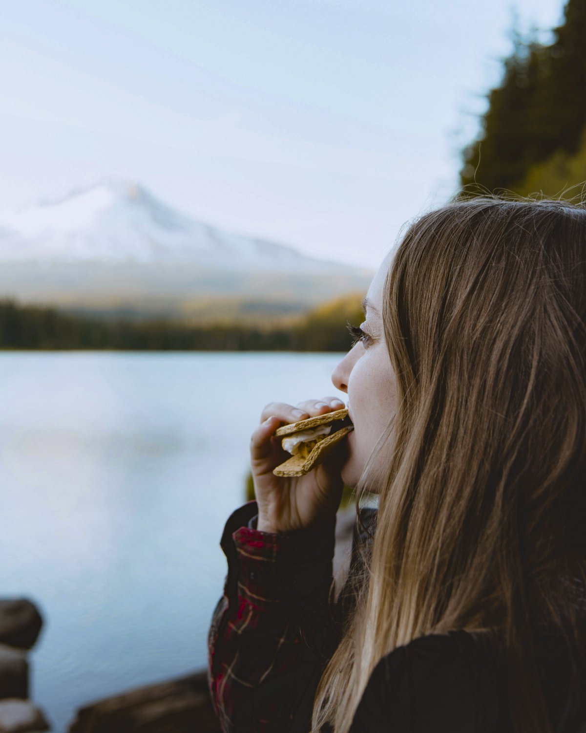 A young woman with long brown hair eating smores, looking out at a lake and ice-capped mountain, likely planning her website