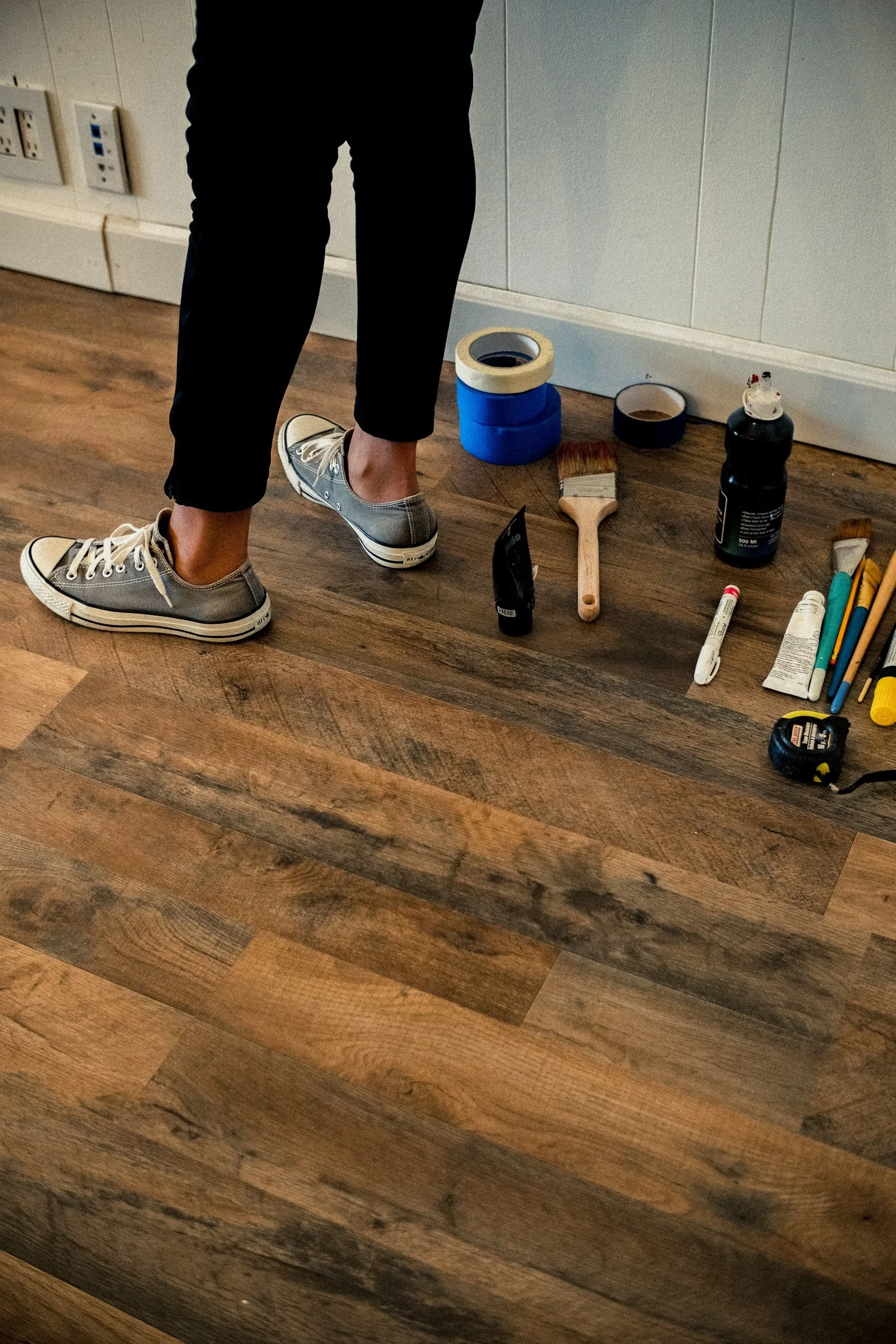 Person standing on a wooden floor with various painting supplies, including brushes, tape, and paint bottles, arranged on the floor nearby.
