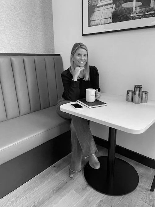 A woman sitting in a booth at a restaurant, smiling and holding a mug, with a smartphone and notepad on the table, and framed artwork on the wall behind her.