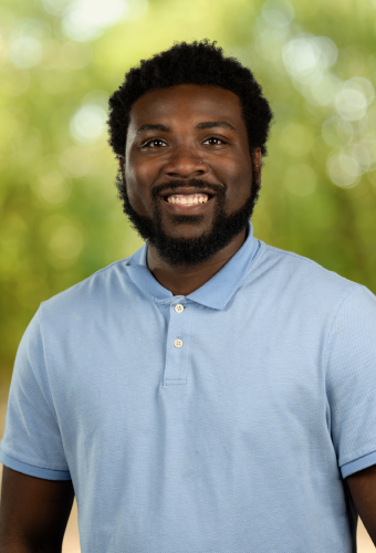 A smiling man with a beard and short curly hair, wearing a light blue polo shirt, standing outdoors with a blurred green background.