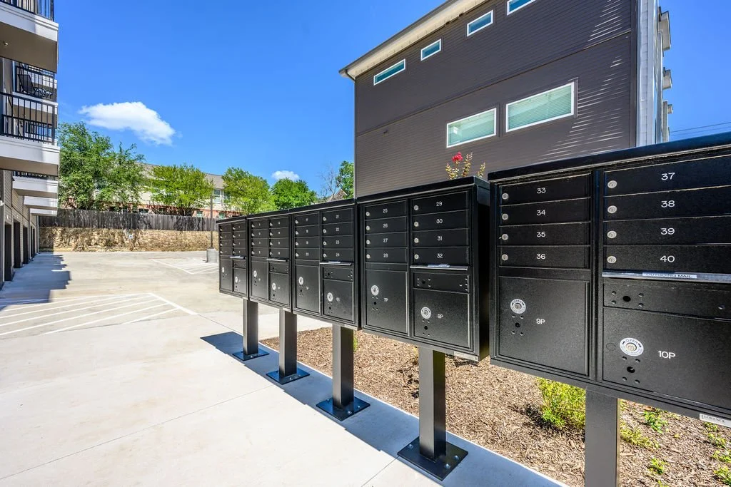 Black mailbox units with numbers on metal posts in an apartment complex parking lot.