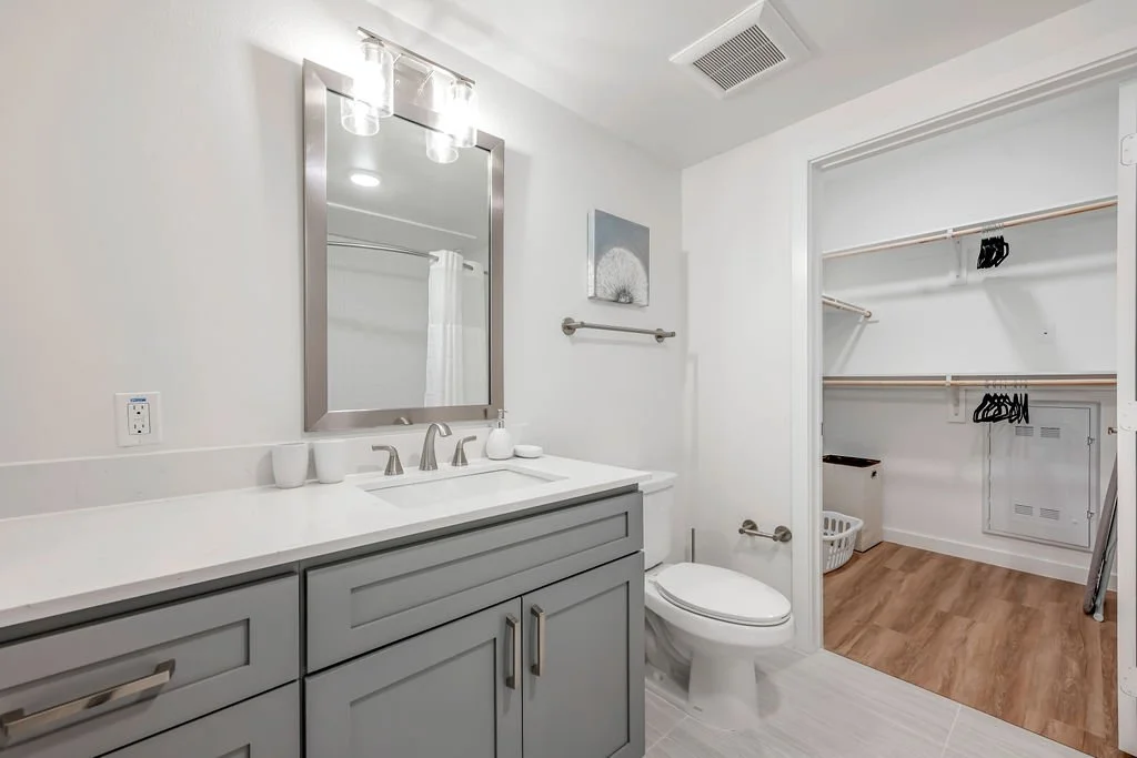 Modern bathroom with a gray vanity, a large mirror, and a white countertop. There are three small cups on the counter, a soap dispenser, and a decorative item. A toilet is next to the vanity. Behind it, there is an open walk-in closet with wooden flo