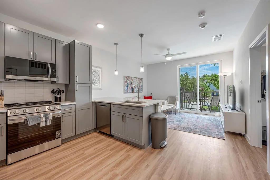 Open-concept kitchen and living area with gray cabinets, stainless steel appliances, a white countertop, a small dining area, a balcony with outdoor furniture, and a TV on a white stand.