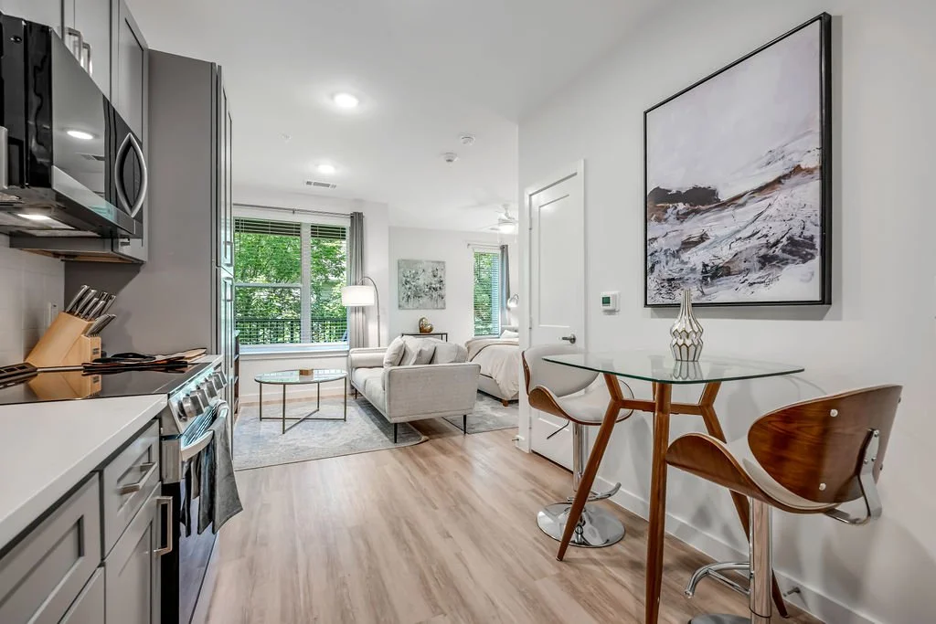 Open-concept living area with kitchen on the left, and a sitting area with a sofa, coffee table, and large windows overlooking trees in the background. To the right, a small dining area with a glass table, two wooden chairs, and a framed abstract pai