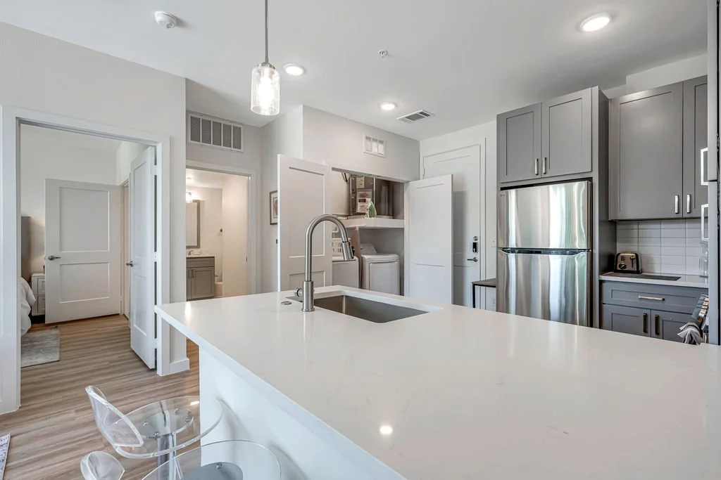 Modern kitchen with gray cabinets, stainless steel refrigerator, white countertop, and a clear acrylic chair.