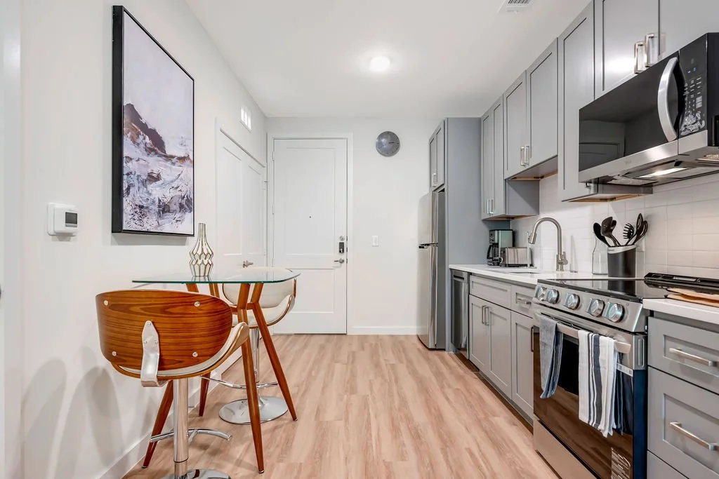 Modern kitchen with gray cabinets, stainless steel appliances, a small glass dining table with two wooden chairs, hardwood floors, and decorative wall art.
