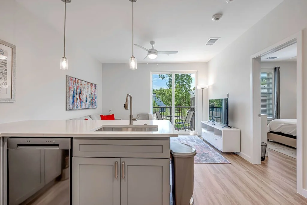 Living room with white walls, a sliding glass door leading to a balcony with outdoor chairs, a TV on a white stand, and a doorway leading to a bedroom with a bed and window.