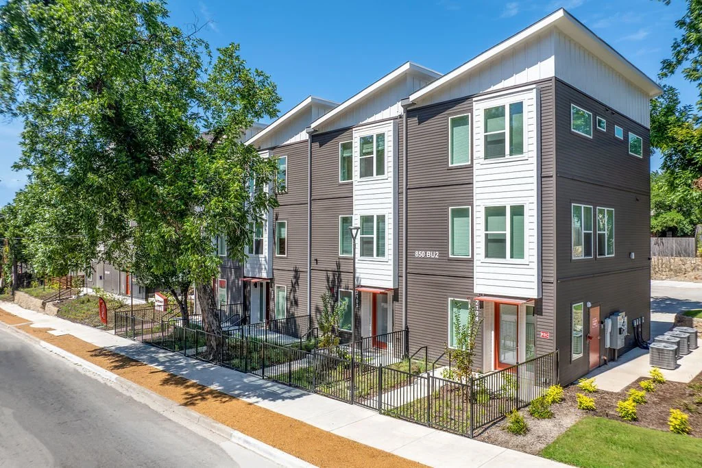 Modern multi-family residential building with brown and white siding, large windows, and small front yards with fencing, trees, and landscaping along a sidewalk under a clear blue sky.