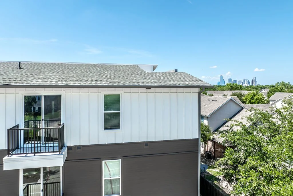 View of a suburban residential area with a white multi-story building and a balcony, with trees and neighboring houses, and a city skyline in the background under a clear blue sky.