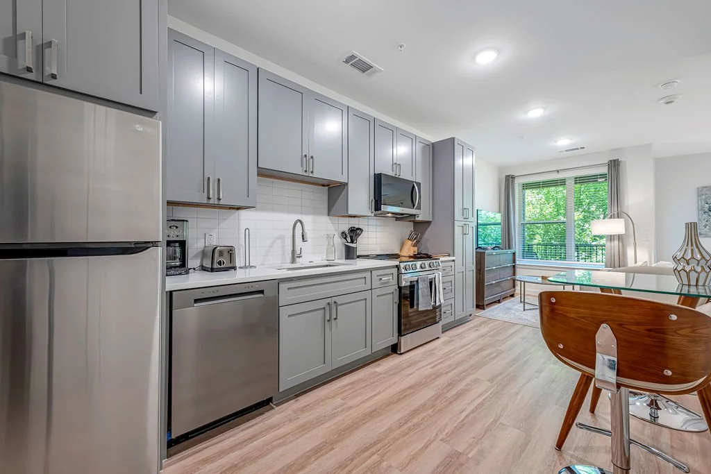 Modern kitchen and dining area with gray cabinets, stainless steel appliances, and a glass dining table with wooden chairs.
