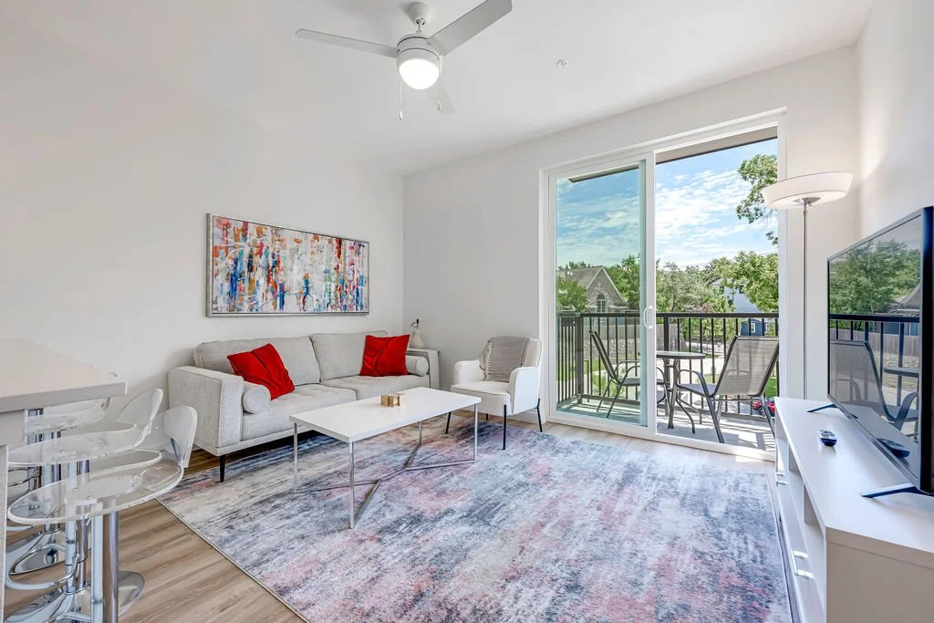 Bright living room with a white sofa, red cushions, abstract painting, sliding glass door leading to balcony, outdoor table and chairs, TV on white stand, and ceiling fan.