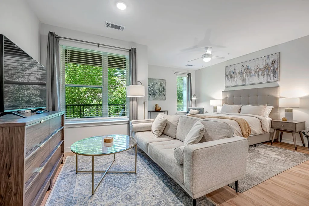 Modern bedroom with large window, light-colored bed, beige sofa, wooden dresser, round glass coffee table, art on walls, and leafy green view outside.