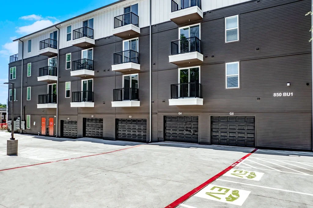 Front view of a multi-story residential building with parking spaces in the foreground, including numbered parking spots and garage doors.