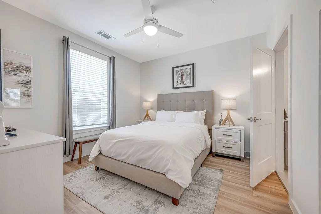 A cozy bedroom with a large bed, twin nightstands with lamps, a window with curtains, and a ceiling fan, decorated in neutral tones.