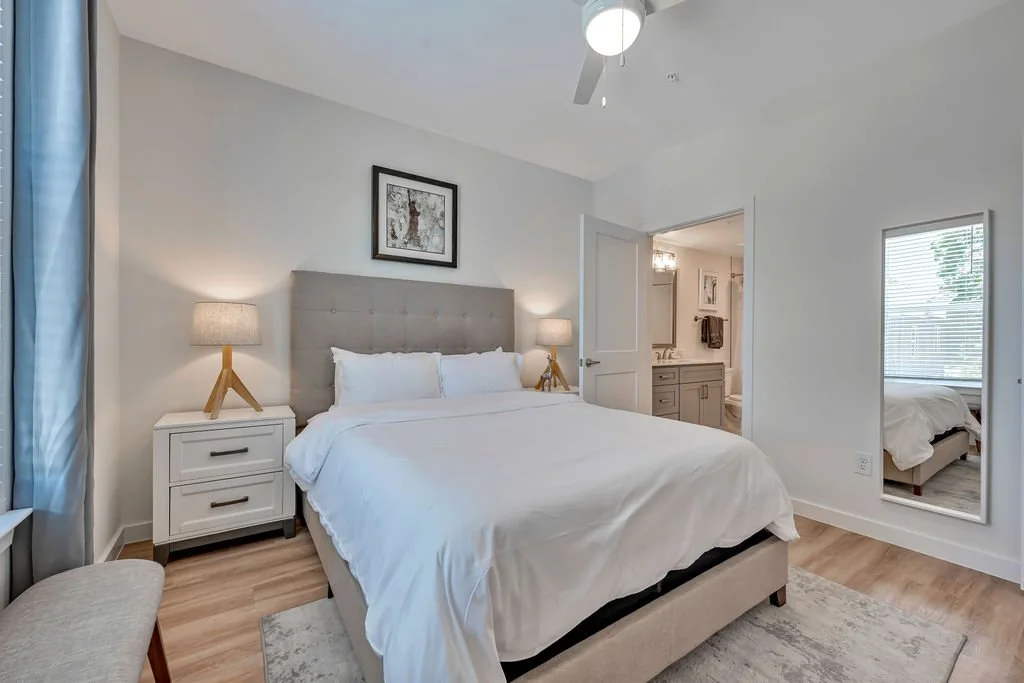 Photo of a modern bedroom with a large bed, white bedding, grey tufted headboard, two white nightstands with lamps, a framed wall art above the headboard, and a mirror on the right wall showing part of a bed in the adjacent room. There is a ceiling f