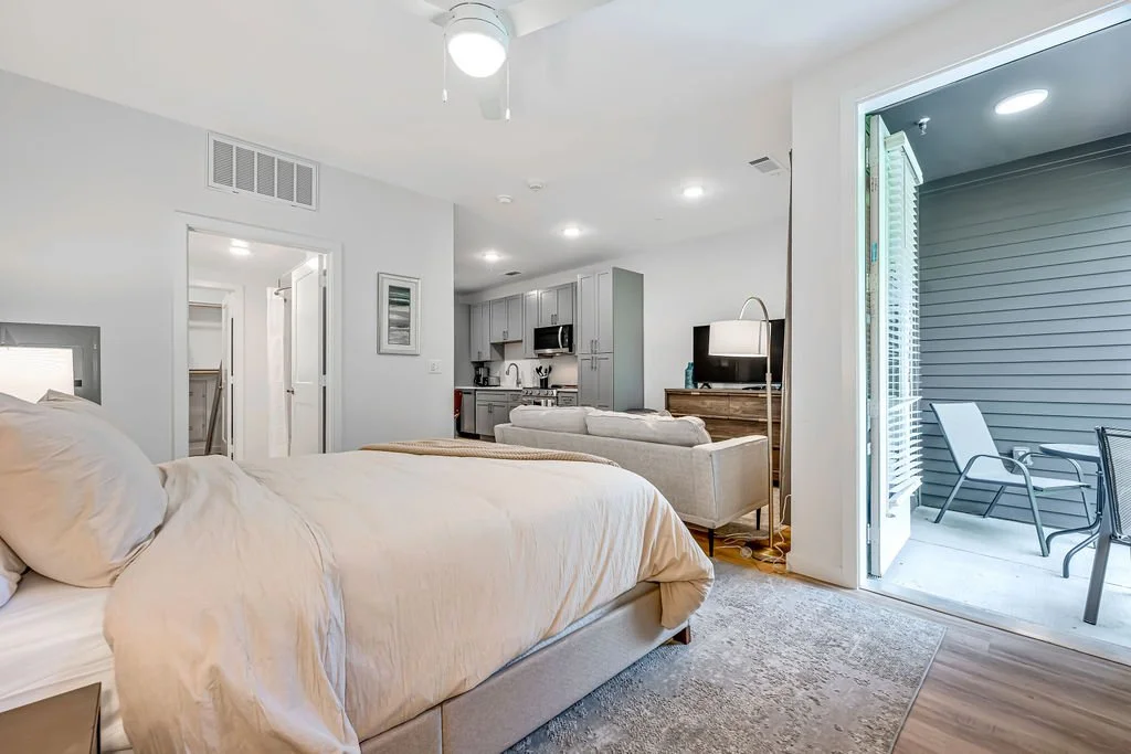 Bright bedroom with a sliding glass door leading to a small outdoor patio with chairs, an adjoining living area with a sofa, TV, and kitchen in the background.