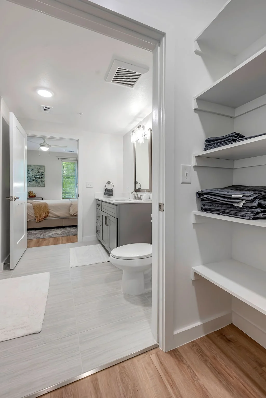 Bathroom with gray vanity, toilet, mirror, and shelves with folded towels, view into bedroom with bed and window.