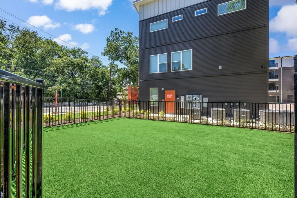 Empty outdoor area with artificial green turf, black metal fence, and modern building with windows and utility boxes in the background.