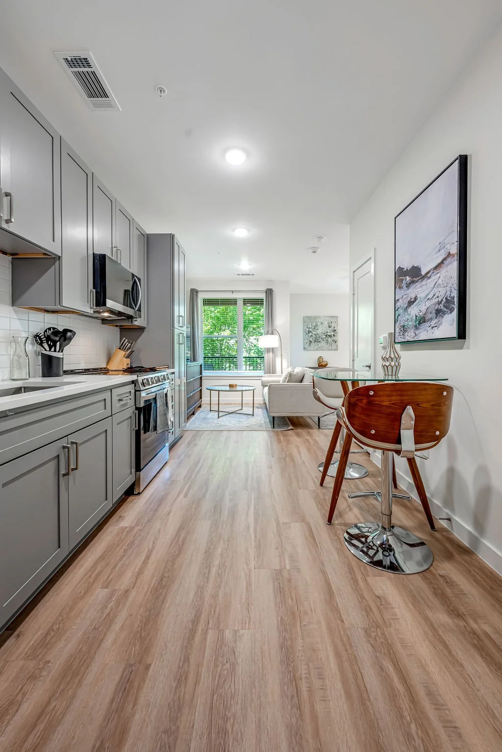 Interior view of a modern apartment with a kitchen on the left, a living area with a sofa and window in the background, and a dining table with a wooden chair on the right.
