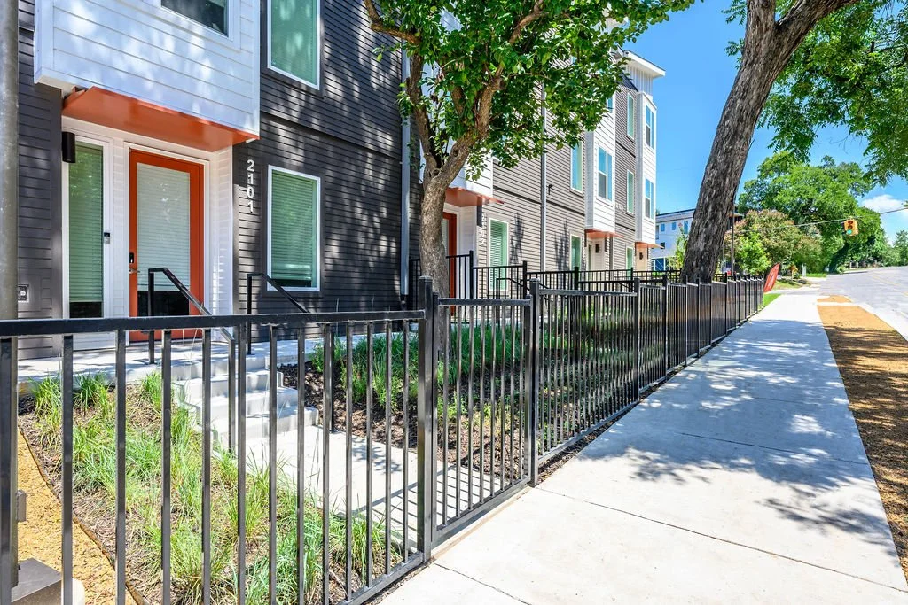 Exterior view of modern multi-story residential buildings with black and white siding, orange door frames, and small front yards enclosed by black metal fences. Sidewalk runs alongside the buildings with trees providing shade.