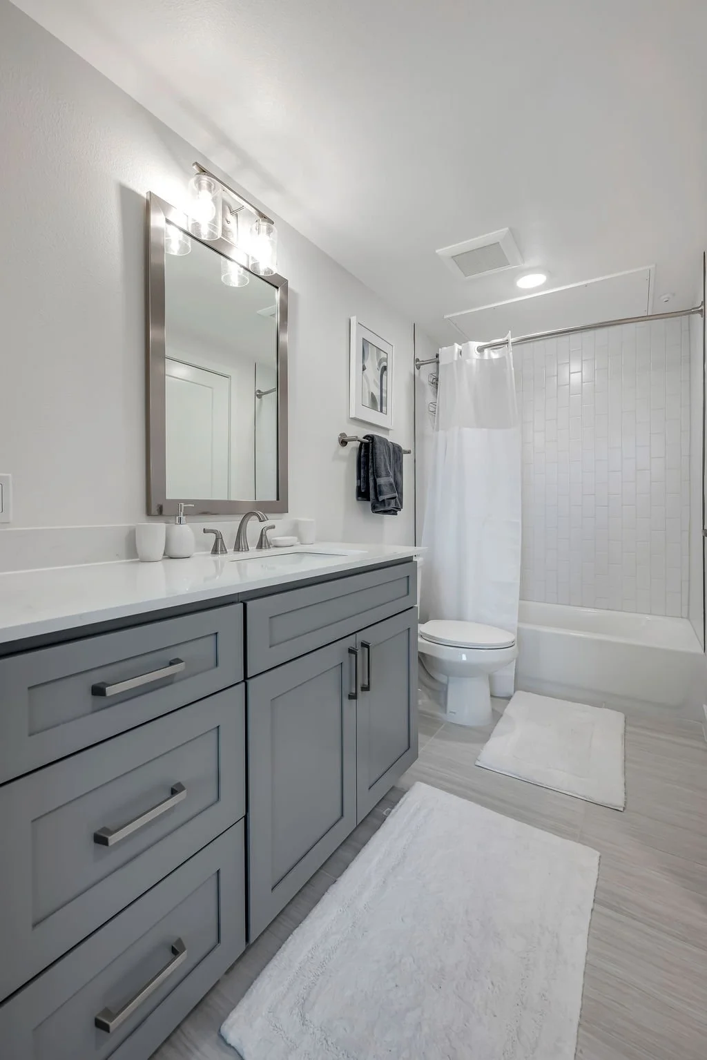 Modern bathroom with gray vanity, white countertop, large mirror, white towels, white bathtub with shower curtain, tiled shower area, toilet, framed artwork, ceiling vent, LED ceiling light, light wood flooring, white bath mats.