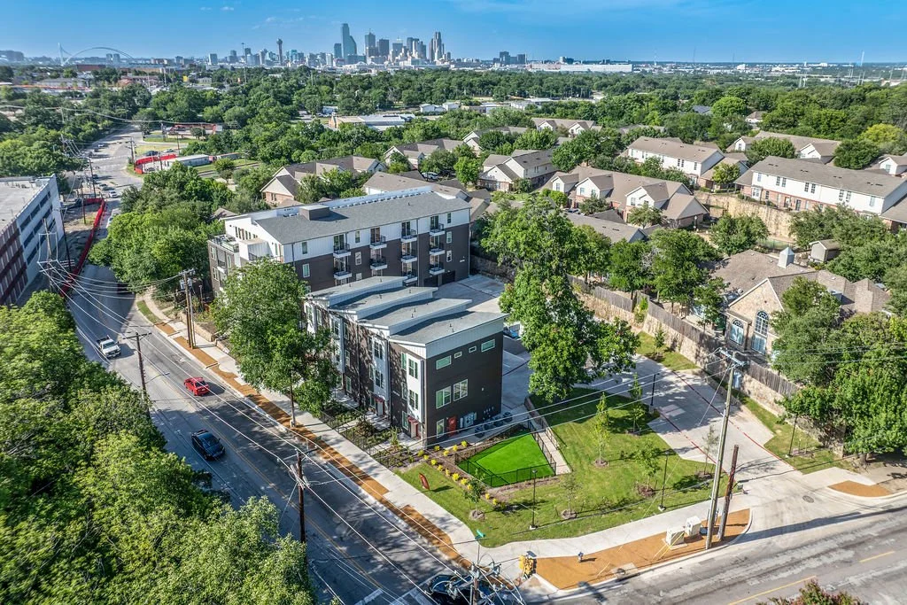Aerial view of a suburban neighborhood with single-family homes, a modern apartment building, and a city skyline in the background under a clear blue sky.