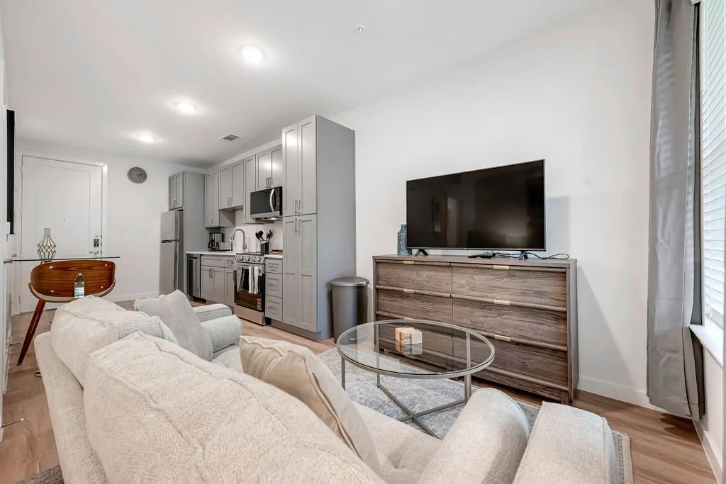 Living room with a beige sofa, a glass coffee table, a wooden TV stand with a large flat-screen TV, a gray kitchen with appliances, and a round dining table with a wooden chair.