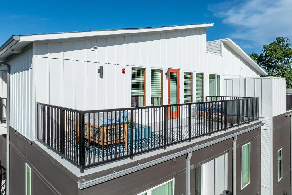 Upper-floor balcony with outdoor furniture, black railing, white exterior walls, and large windows on a modern apartment building.