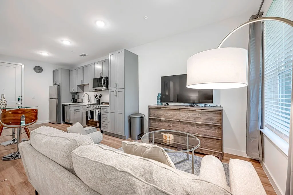 Living room and kitchen area with a beige sofa, glass coffee table, TV on a wooden dresser, kitchen with gray cabinets, stainless steel appliances, and a dining area with a wooden table and chairs.