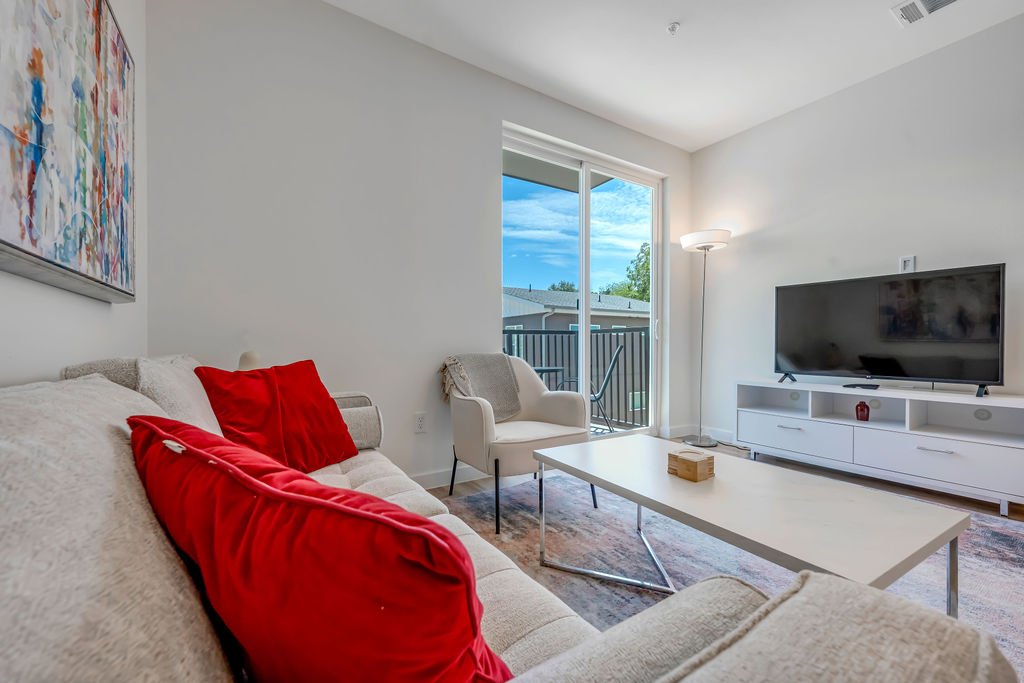 Bright living room with a large beige sofa and red cushions, white armchair, white coffee table, and a flat-screen TV on a white TV stand, with a sliding glass door leading to a balcony and an outdoor view of a neighboring building and blue sky.