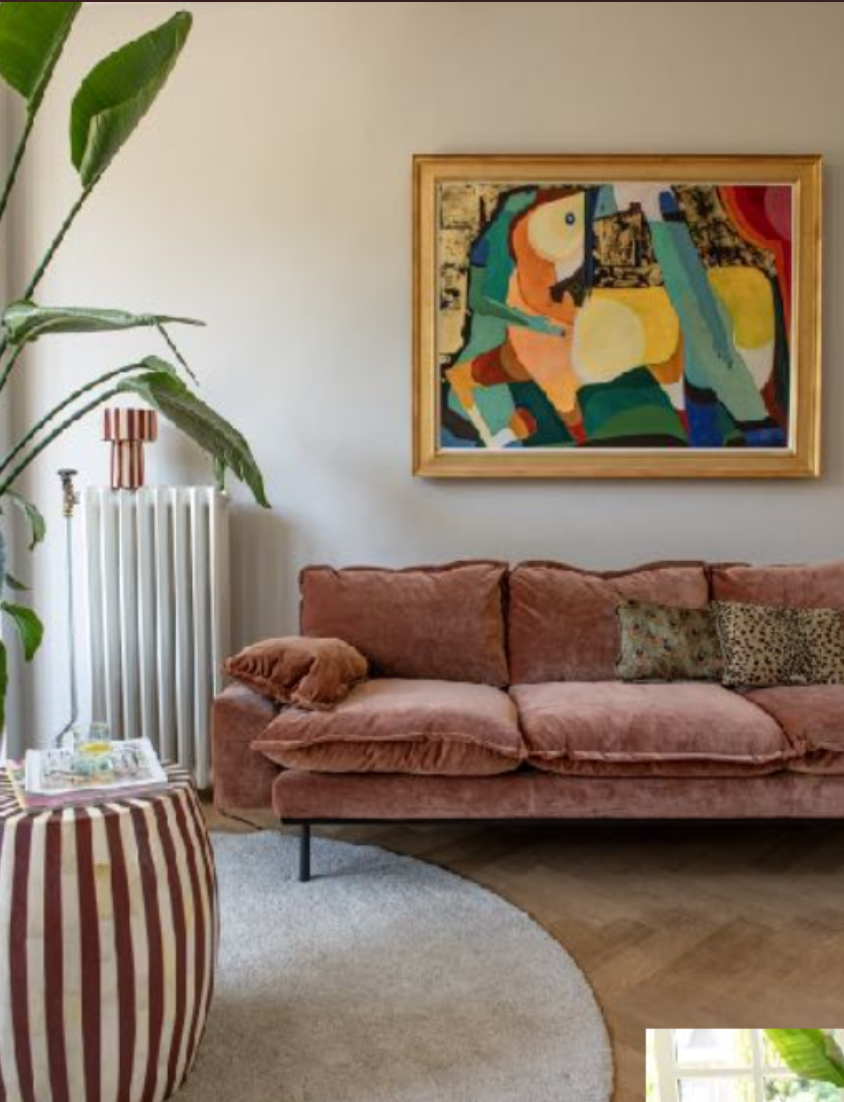 Woonkamer met een roze bank, een kleurrijk abstract kunstwerk aan de muur, een grote groene plant, een lamp met roze en witte strepen, een ronde side table met een wit dienblad, een witte radiator, een beige tapijt en een houten vloer.